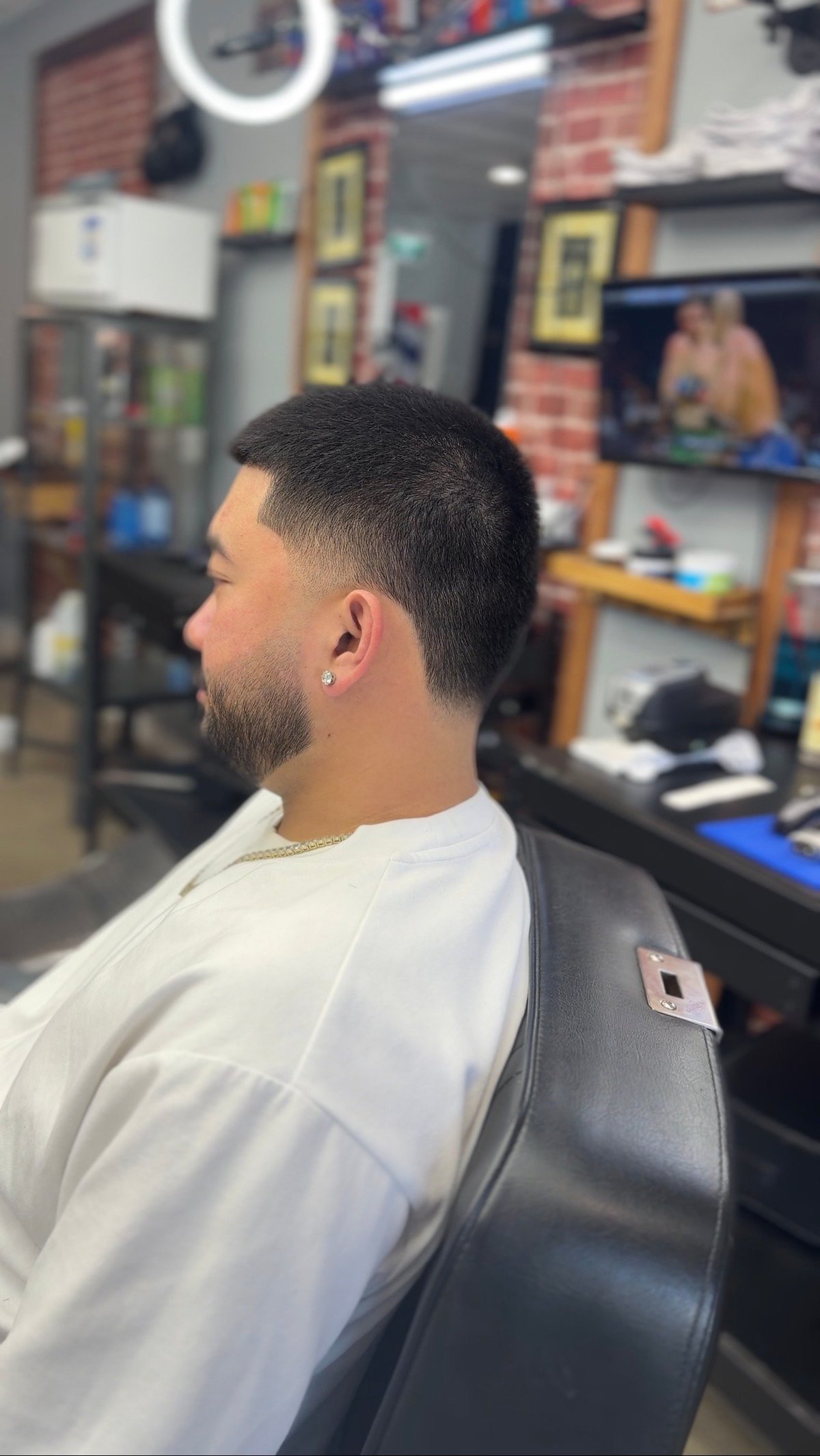 Man with short dark hair and beard sitting in barber chair in salon, side profile view