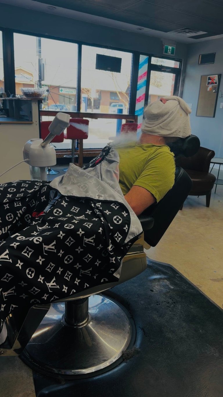 Person reclined in barber chair wearing yellow shirt and beige head covering, with salon interior and windows visible in background