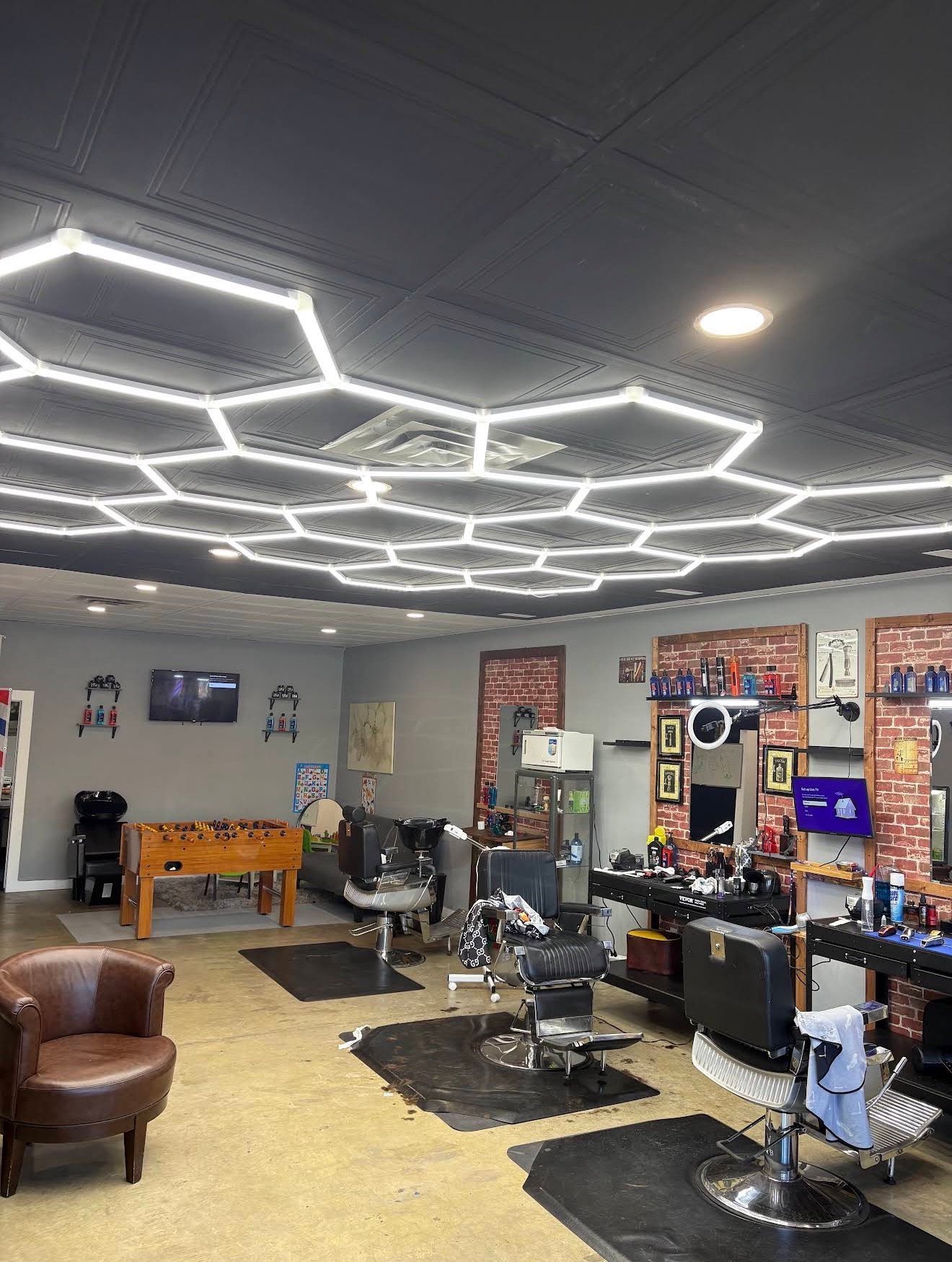 Modern barbershop interior with neon ceiling lights, barber chairs, exposed brick wall, pool table, and lounge seating area
