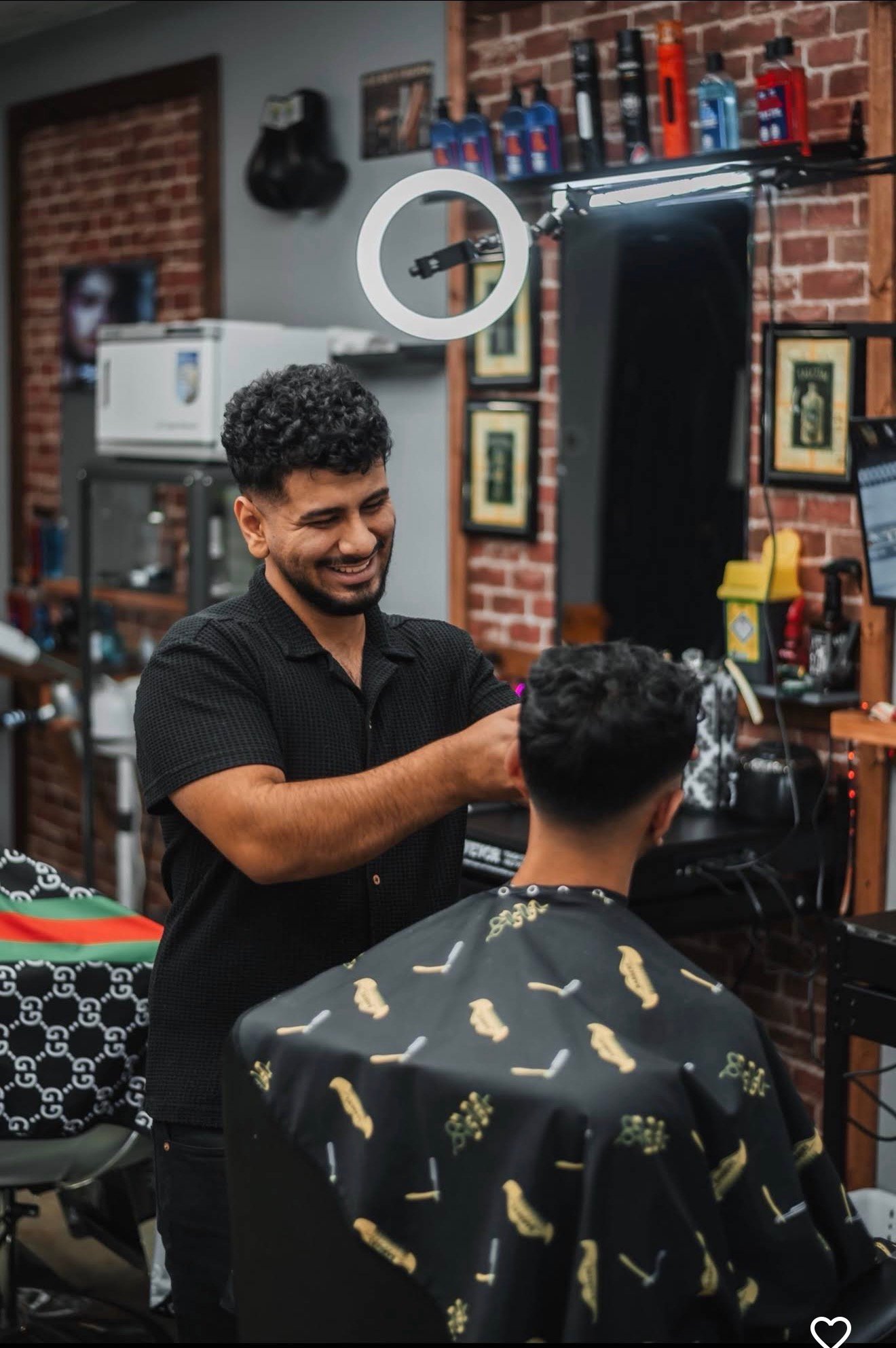 Barber cutting clients hair in a professional salon with exposed brick walls and equipment visible in the background